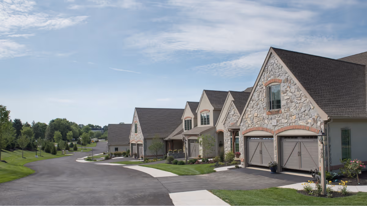 Row of stone and brick residential buildings with garages along a curved paved road, surrounded by well-maintained green lawns and trees under a partly cloudy sky.