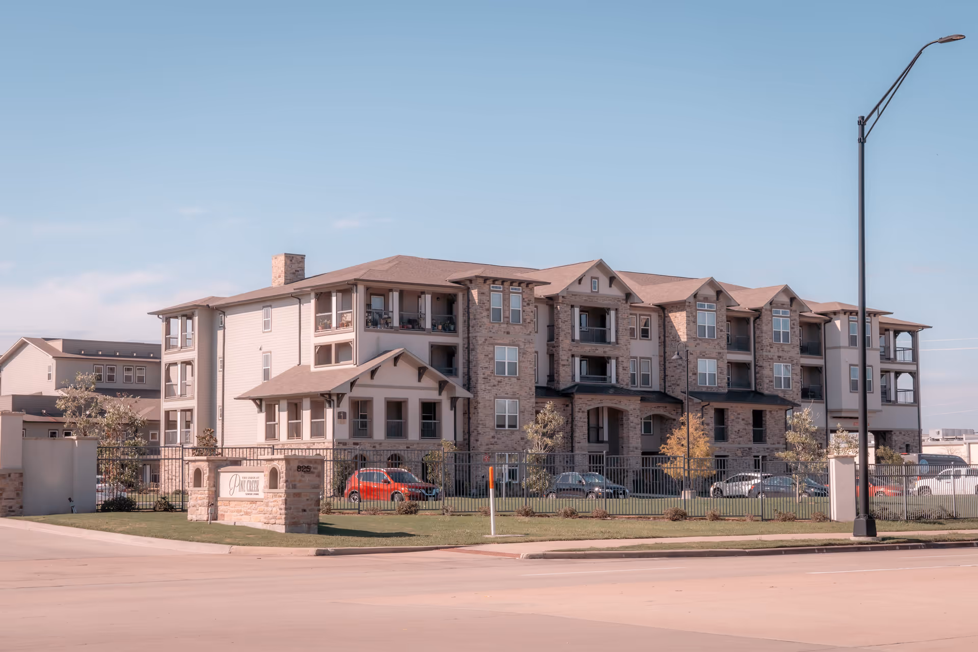Exterior view of a multi-story senior living facility named The Lodge at Pine Creek, featuring a combination of brick and siding, balconies, a fenced lawn area, parked cars, and a clear blue sky.