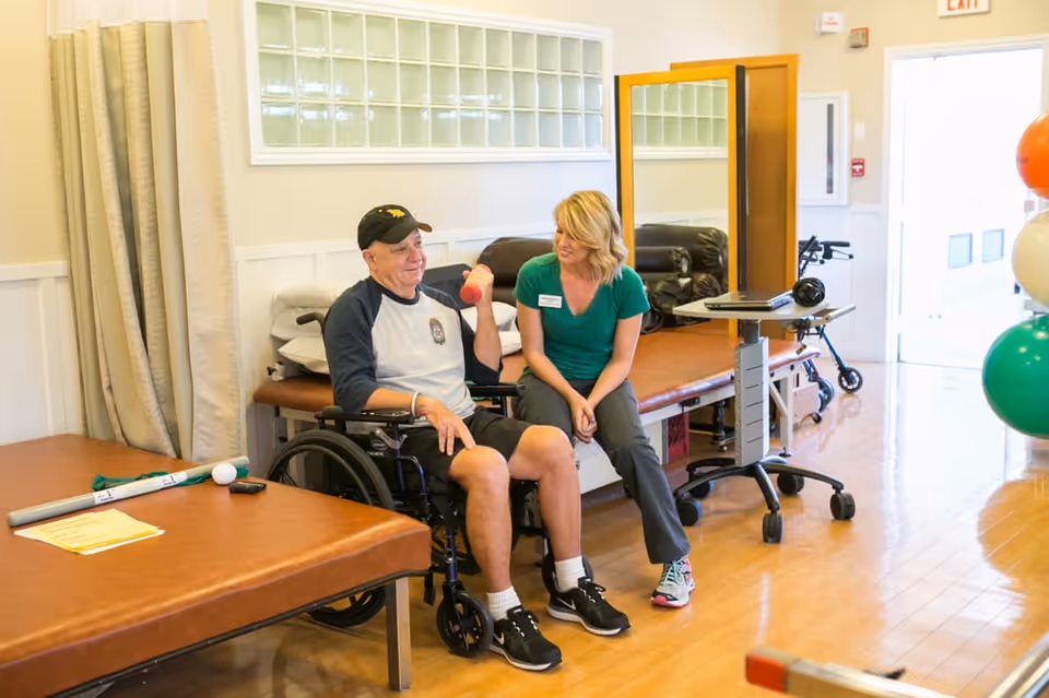 A man in a wheelchair lifting a small dumbbell while a woman, possibly a caregiver or therapist, sits beside him smiling in a rehabilitation or therapy room with exercise equipment and a large mirror.