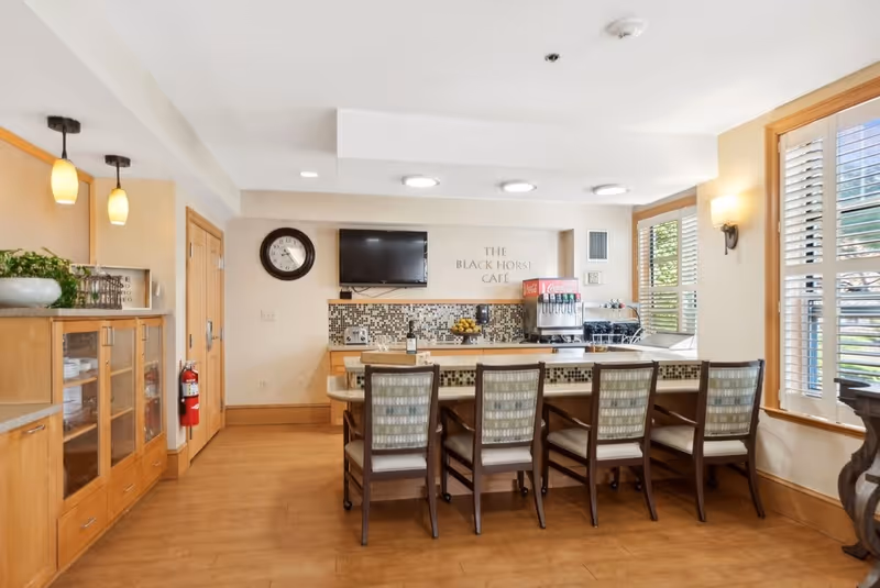 Interior view of The Black Horse Cafe featuring a counter with six chairs, a wall-mounted TV, a clock, a beverage dispenser, and wooden cabinets with dishes. The room has large windows with white shutters allowing natural light to enter.