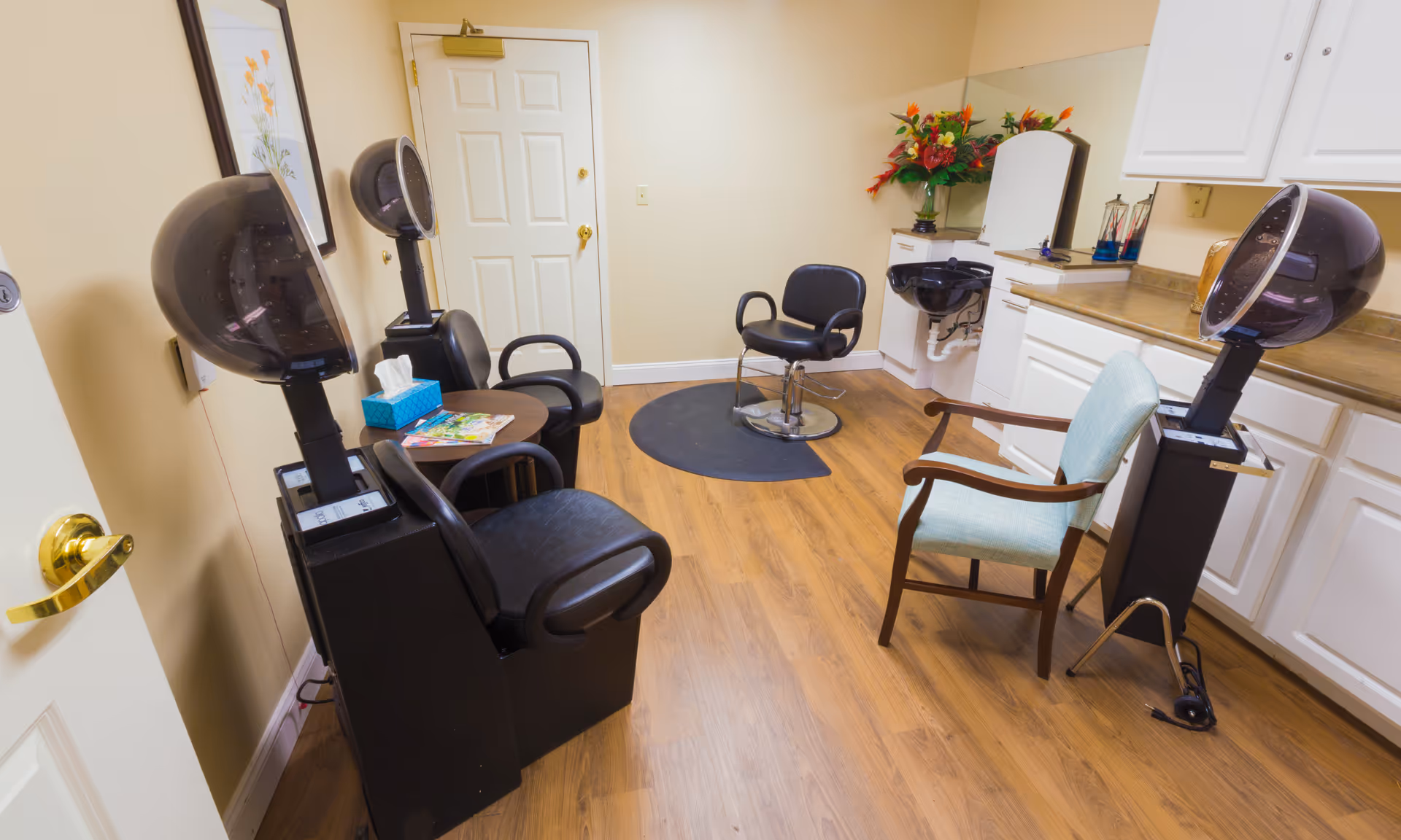A small salon-style room with hair dryers, styling chairs, a sink, cabinets and a waiting chair.