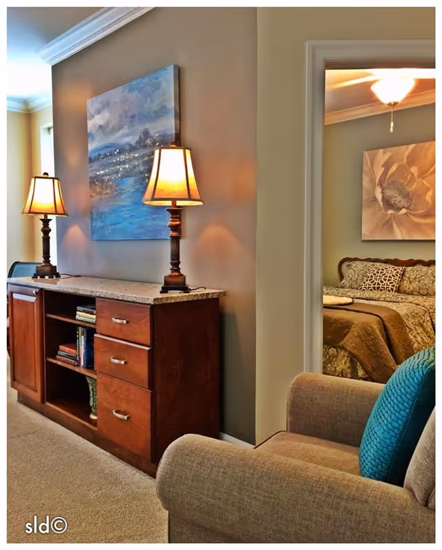 Interior view of a senior living facility showing a wooden cabinet with two lit table lamps and a painting above it. To the right, there is a partial view of a bedroom with a bed covered in patterned bedding and a large floral artwork on the wall. In the foreground, there is a beige armchair with a blue cushion.