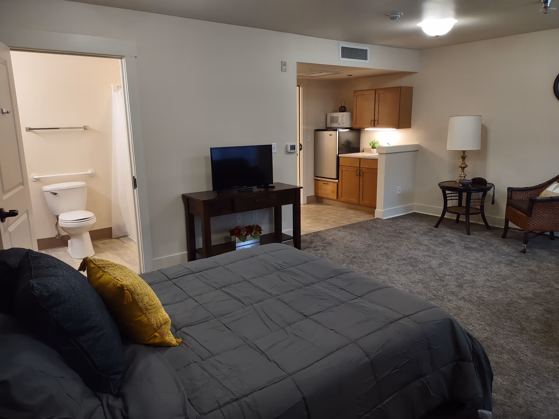 Interior view of a senior living facility room featuring a bed with dark bedding and pillows, a small TV on a wooden stand, an open bathroom door showing a toilet and shower curtain, a kitchenette with wooden cabinets, a mini fridge, and a microwave, and a sitting area with a chair, side table, and lamp.