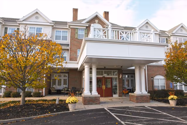 Front entrance of a brick senior living building with a covered portico, white columns, red double doors, and autumn trees.