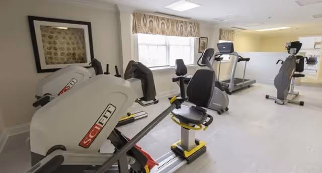 A bright fitness room with exercise equipment including stationary bikes, a rowing machine, and a treadmill. The room has light-colored walls, a large window with patterned curtains, and framed artwork on the wall.