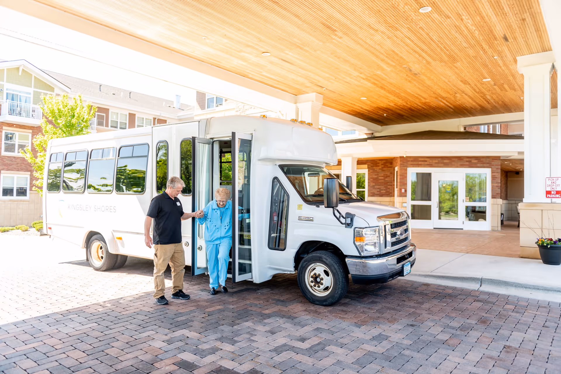 A man assists an elderly woman as she steps off a white shuttle bus outside a senior living facility. The bus has 'Kingsley Shores' written on its side. The area is covered with a wooden ceiling and brick-paved ground, with the facility entrance visible in the background.