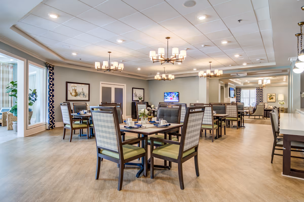 Bright communal dining room with multiple set tables and chairs, chandeliers, and a TV in a senior living facility.