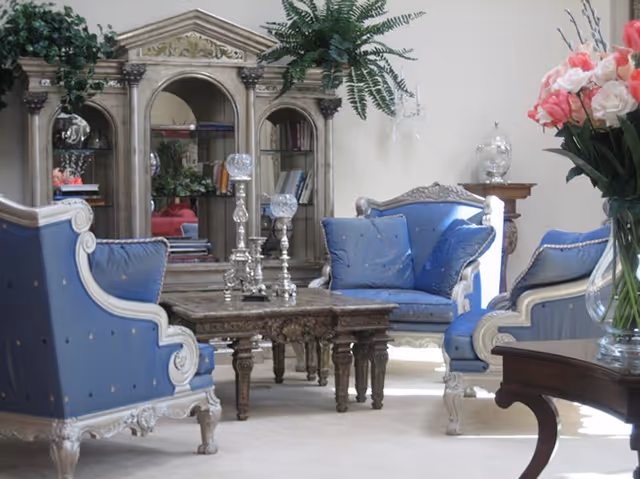 A living room area with ornate blue upholstered armchairs and a matching loveseat surrounding a wooden coffee table. Behind the seating is a large decorative wooden cabinet with a mirror and shelves holding books and plants. A vase with pink and white flowers is visible on the right side.