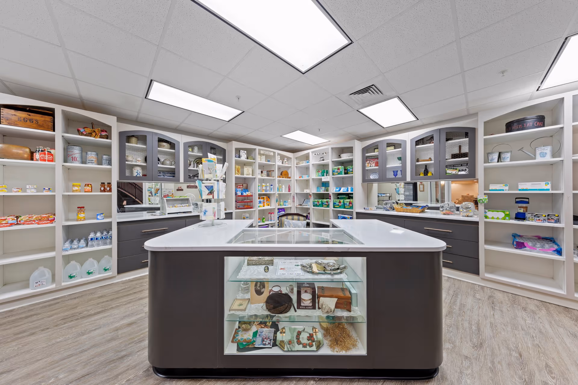 Interior view of a well-lit room with white shelves stocked with various packaged goods, bottled water, and household items. In the center, there is a large island with a glass display case containing decorative items and memorabilia. The room has a clean, organized appearance with a light wood floor and a drop ceiling with fluorescent lights.