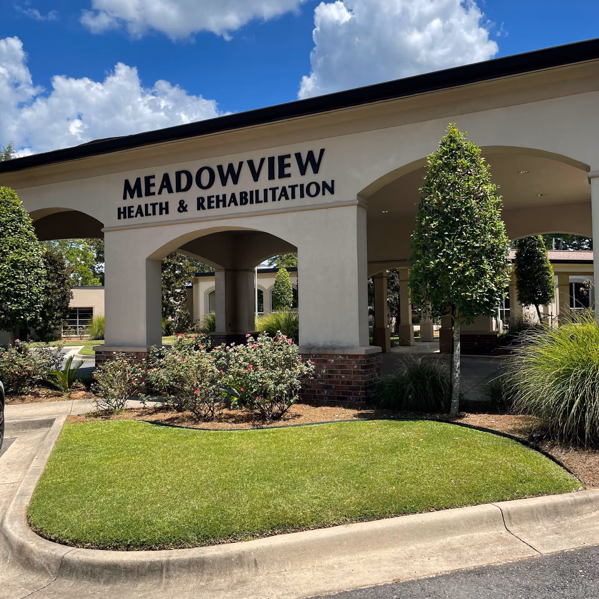 Entrance canopy of Meadowview Health & Rehabilitation Center with manicured lawn, shrubs, and trees under a blue sky.