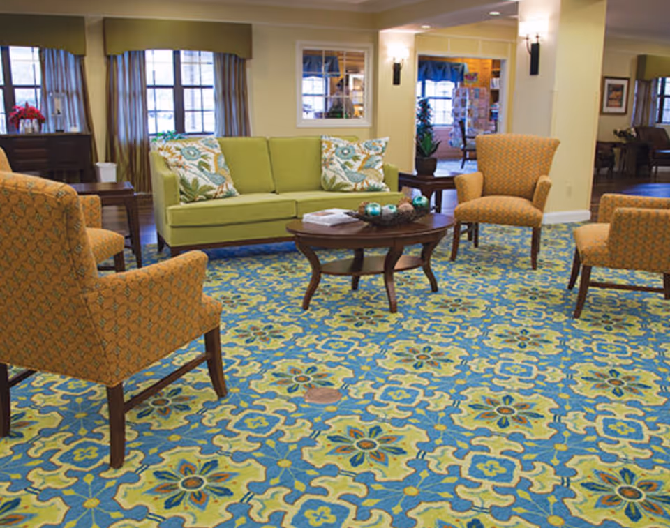 A cozy living room area with a green sofa adorned with floral patterned pillows, surrounded by four orange patterned armchairs. A wooden coffee table with decorative items sits in the center on a colorful blue, green, and yellow patterned carpet. The room has windows with curtains and warm lighting fixtures on the walls.