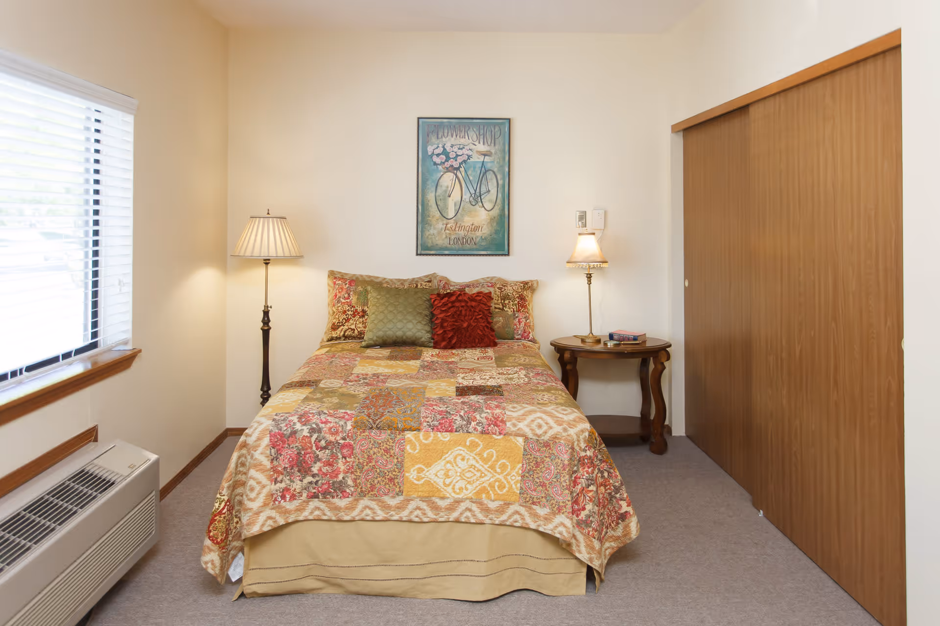 A cozy bedroom with a bed covered in a colorful patchwork quilt and three decorative pillows. There is a floor lamp on the left side and a small wooden side table with a lamp and books on the right. A framed picture of a bicycle with flowers hangs on the wall above the bed. A window with blinds is on the left wall, and a large wooden sliding closet door is on the right.