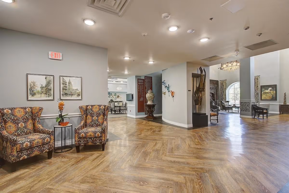 Spacious senior living lobby with patterned armchairs, herringbone wood flooring, and decorative art and seating areas.