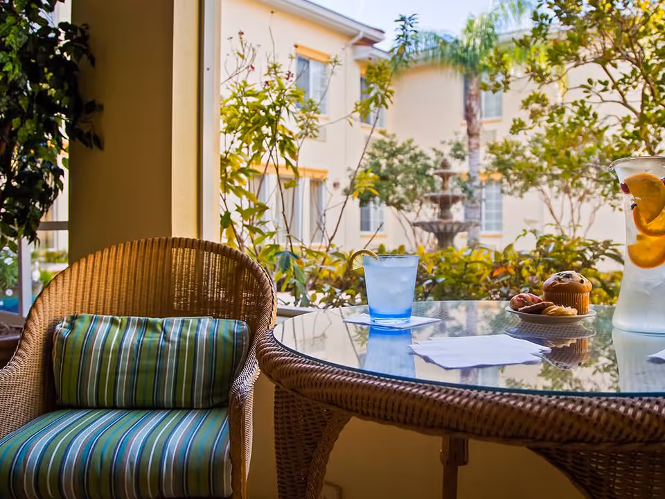 Wicker chair and glass-top table on a covered patio with a glass of water, a muffin and a pitcher, overlooking a leafy courtyard and fountain.