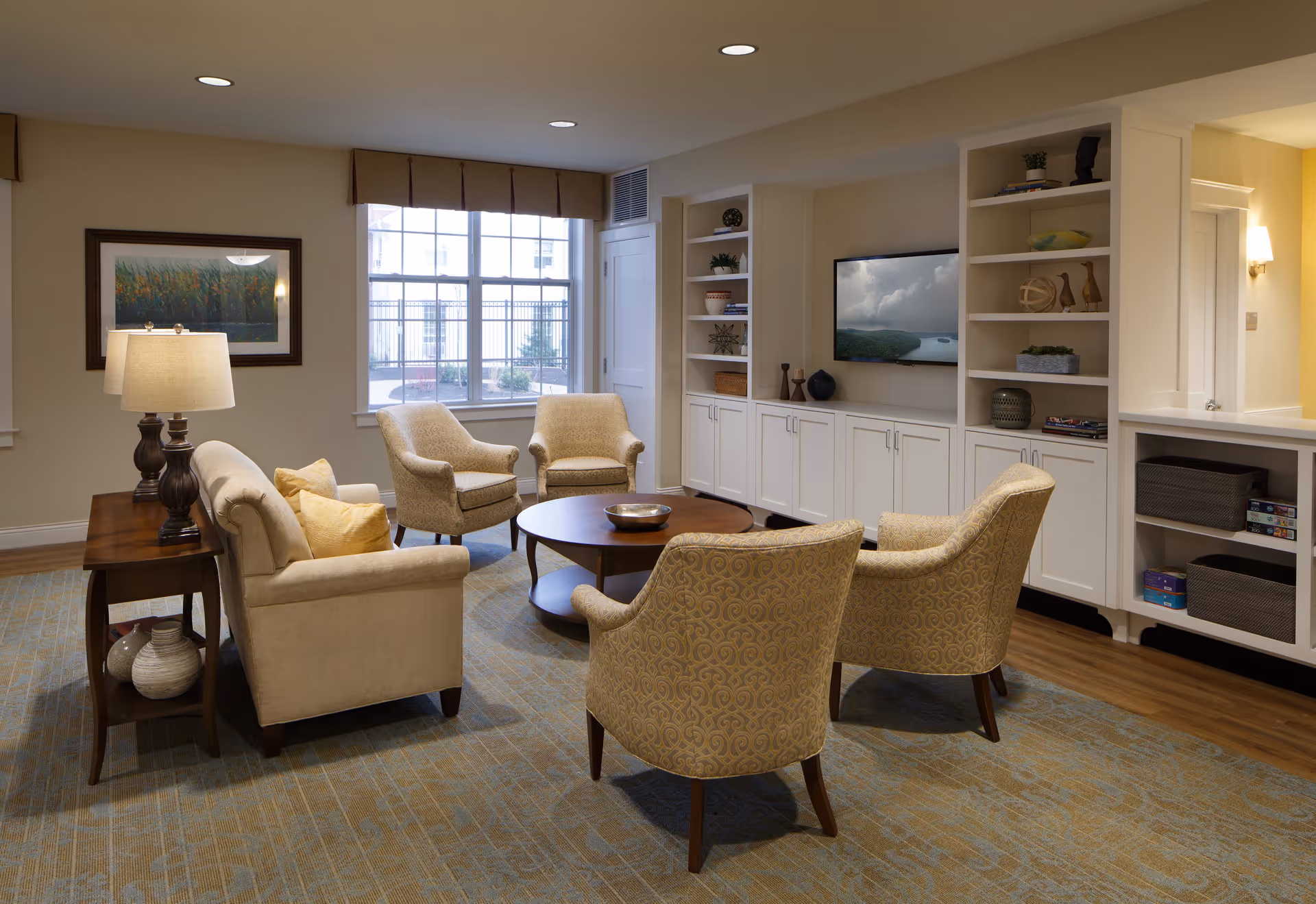 A cozy living room with beige armchairs and a sofa arranged around a round wooden coffee table. The room features built-in white cabinetry with shelves displaying decorative items and a flat-screen TV mounted on the wall. A large window with a brown valance lets in natural light, and a table lamp sits on a wooden side table next to the sofa. The floor is covered with a patterned carpet.