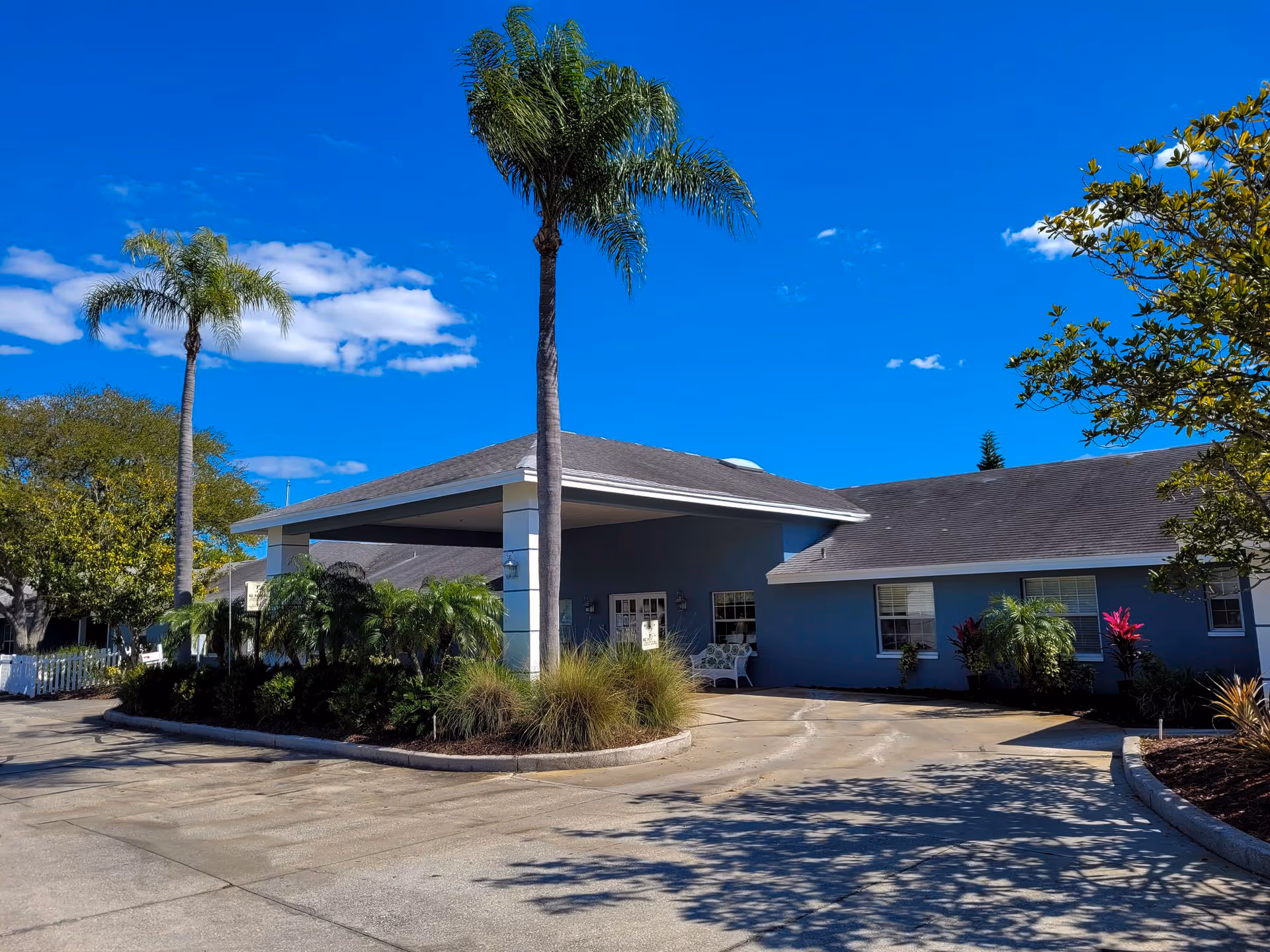 Exterior view of a single-story building with a covered entrance, surrounded by palm trees and landscaping under a clear blue sky.