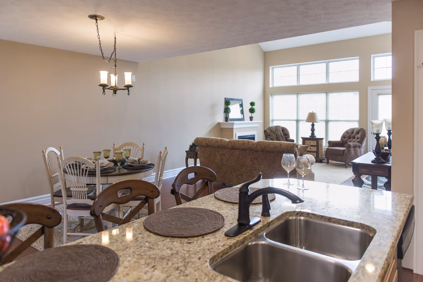 View of an open-concept living space featuring a kitchen island with a double sink and two wine glasses, a dining area with a round table set for four, and a living room with a sofa, armchairs, a fireplace, and large windows allowing natural light.