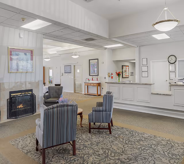 Bright assisted-living reception lobby with striped armchairs around a small table, a fireplace, and a front desk.