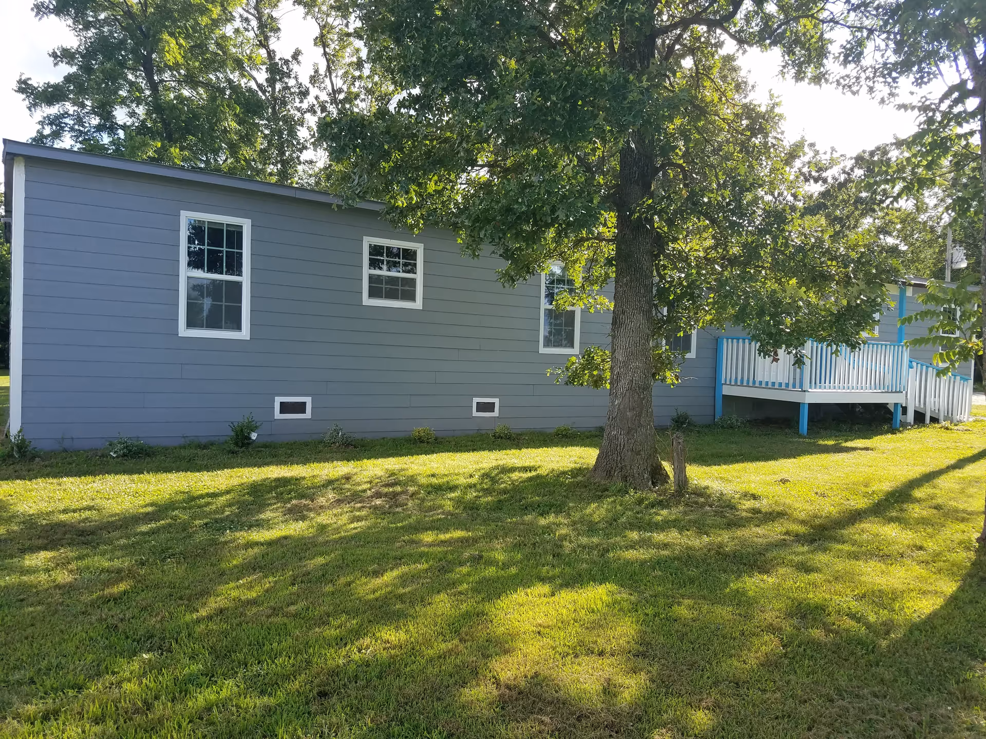 Side view of a single-story gray building with white-trimmed windows and a small porch with blue railings, surrounded by green grass and a large tree casting shadows on the lawn.