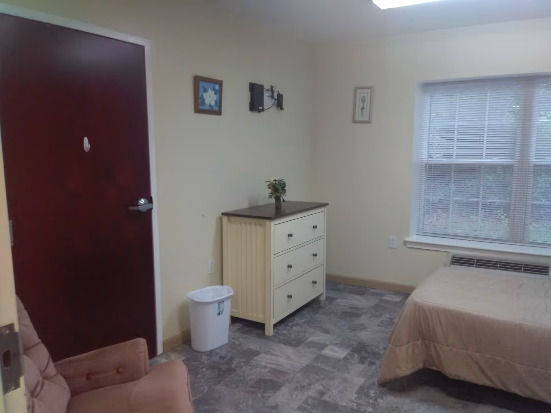 A small bedroom with a beige bedspread on a bed, a white dresser with a small plant on top, a pink cushioned chair, a dark wooden door, two framed pictures on the wall, a window with blinds, and a white trash bin on a tiled floor.