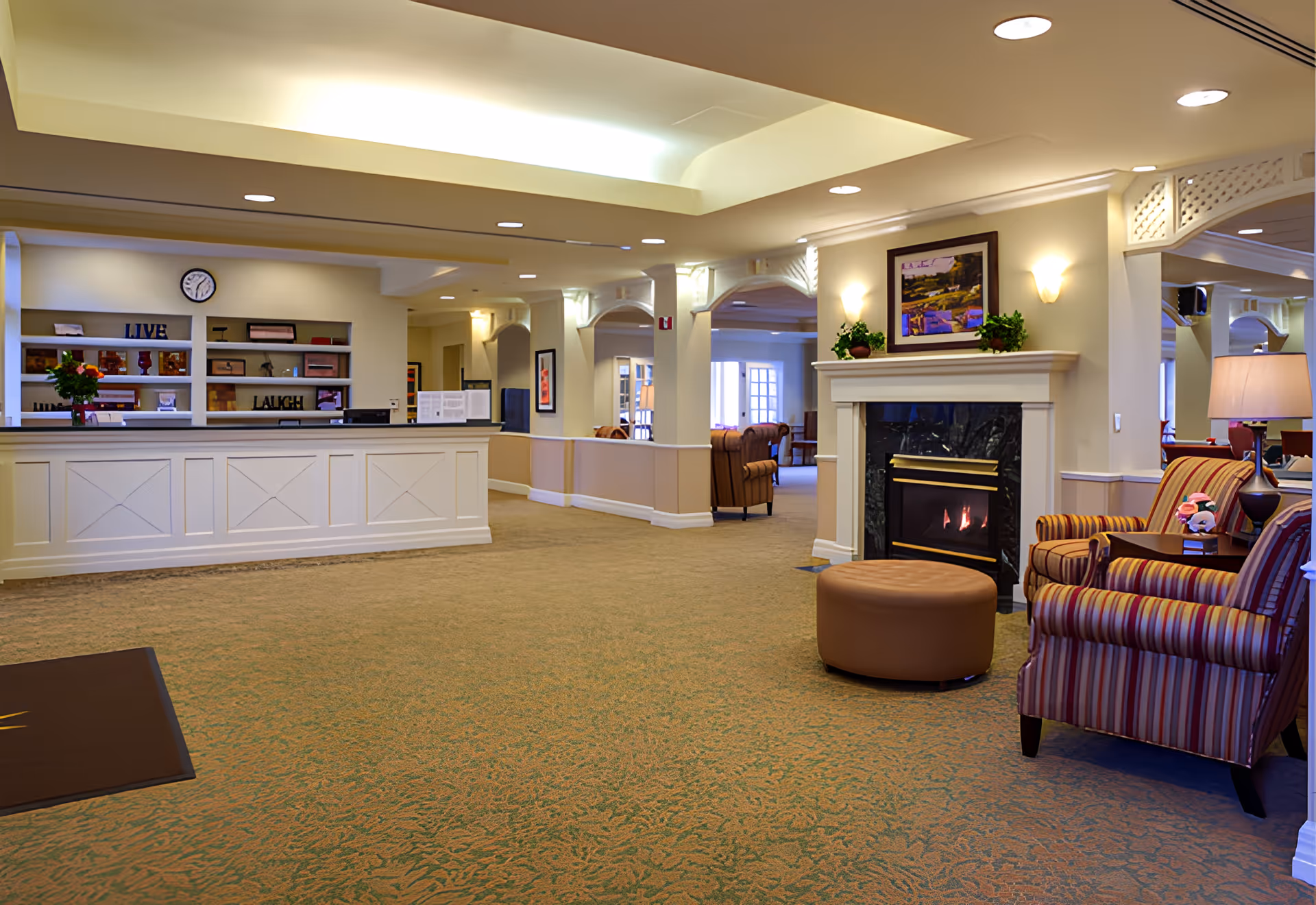 A spacious and well-lit senior living facility common area featuring a reception desk with shelves behind it, a fireplace with a painting and plants on the mantel, and comfortable striped armchairs with a round ottoman. The carpet is patterned, and the ceiling has recessed lighting and a cove light feature.
