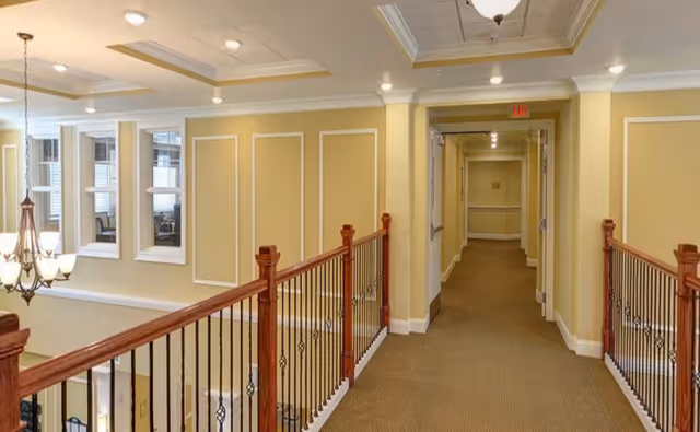 Interior hallway of a senior living facility with beige walls, carpeted floor, wooden railing with black metal balusters, and ceiling lights. There are windows on the left side overlooking a lower floor and a chandelier hanging from the ceiling.