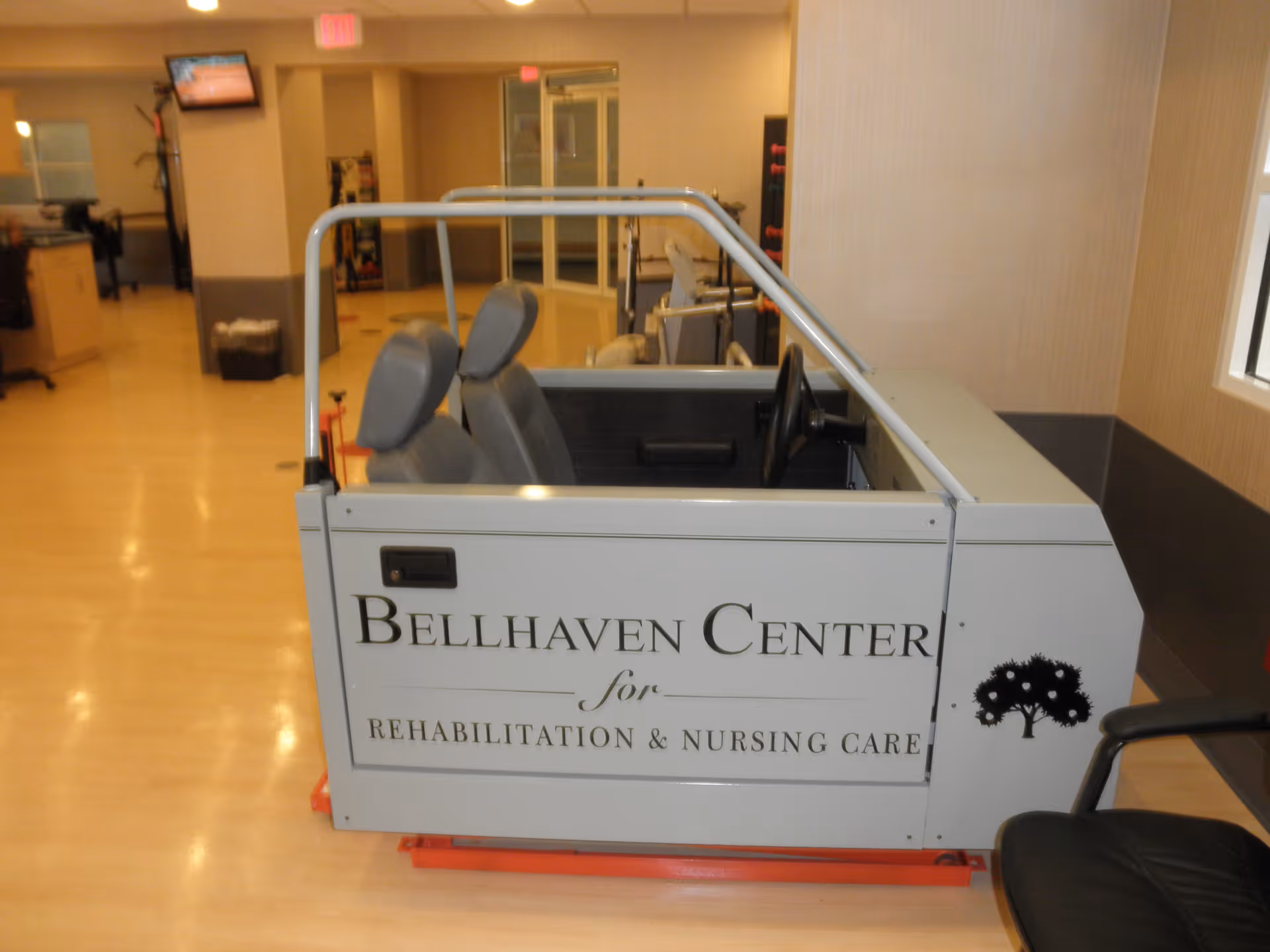 A partial mock-up of a car interior with two gray seats and a steering wheel, labeled 'Bellhaven Center for Rehabilitation & Nursing Care,' placed indoors in a facility with light-colored flooring and walls.