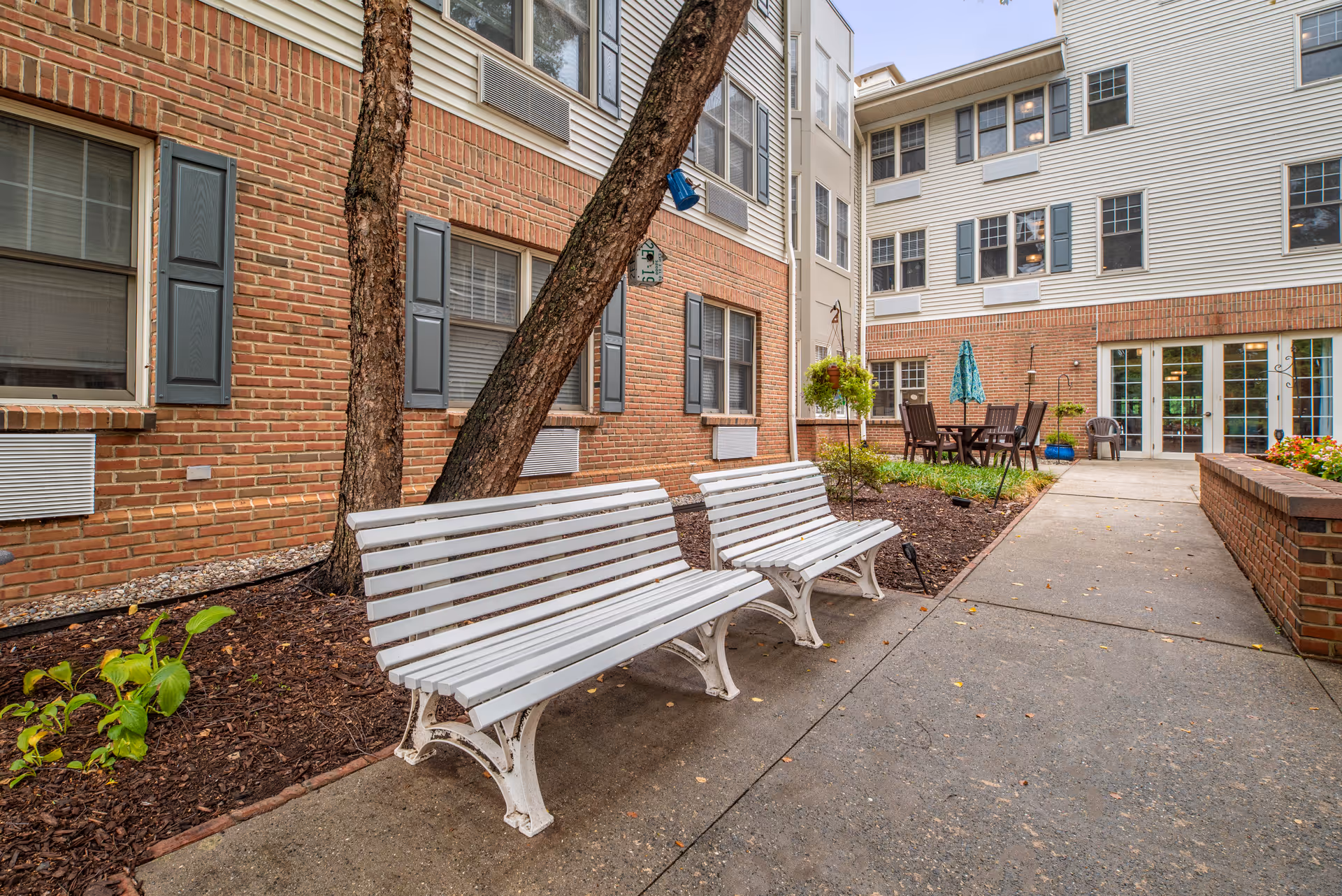 Outdoor courtyard area at Brookdale Monroe featuring two white benches, a brick building with multiple windows, a concrete pathway, and a seating area with a table and umbrella surrounded by plants and greenery.