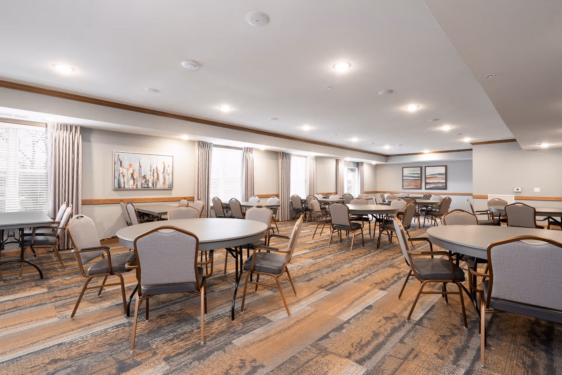 Spacious common dining/activity room with multiple round tables and chairs in a senior living facility.