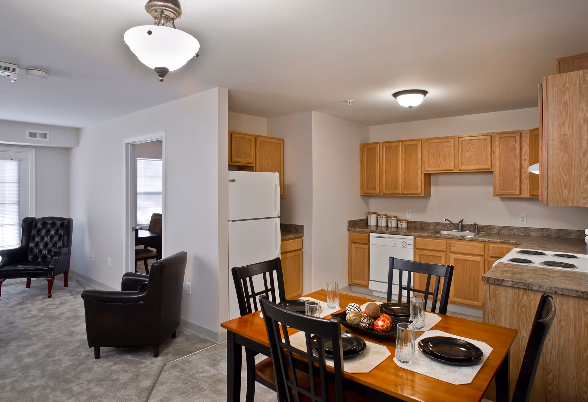 Interior view of a retirement community apartment showing a kitchen with wooden cabinets, a white refrigerator, dishwasher, and stove. In front of the kitchen is a wooden dining table set with four black chairs, black plates, glasses, and decorative centerpiece balls. To the left, there is a living area with two black leather armchairs and a window with blinds.