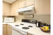 A compact kitchen with light wood cabinets, a white electric stove with four burners, a white microwave on the countertop, a white range hood, and a bowl of fruit including bananas on the counter.