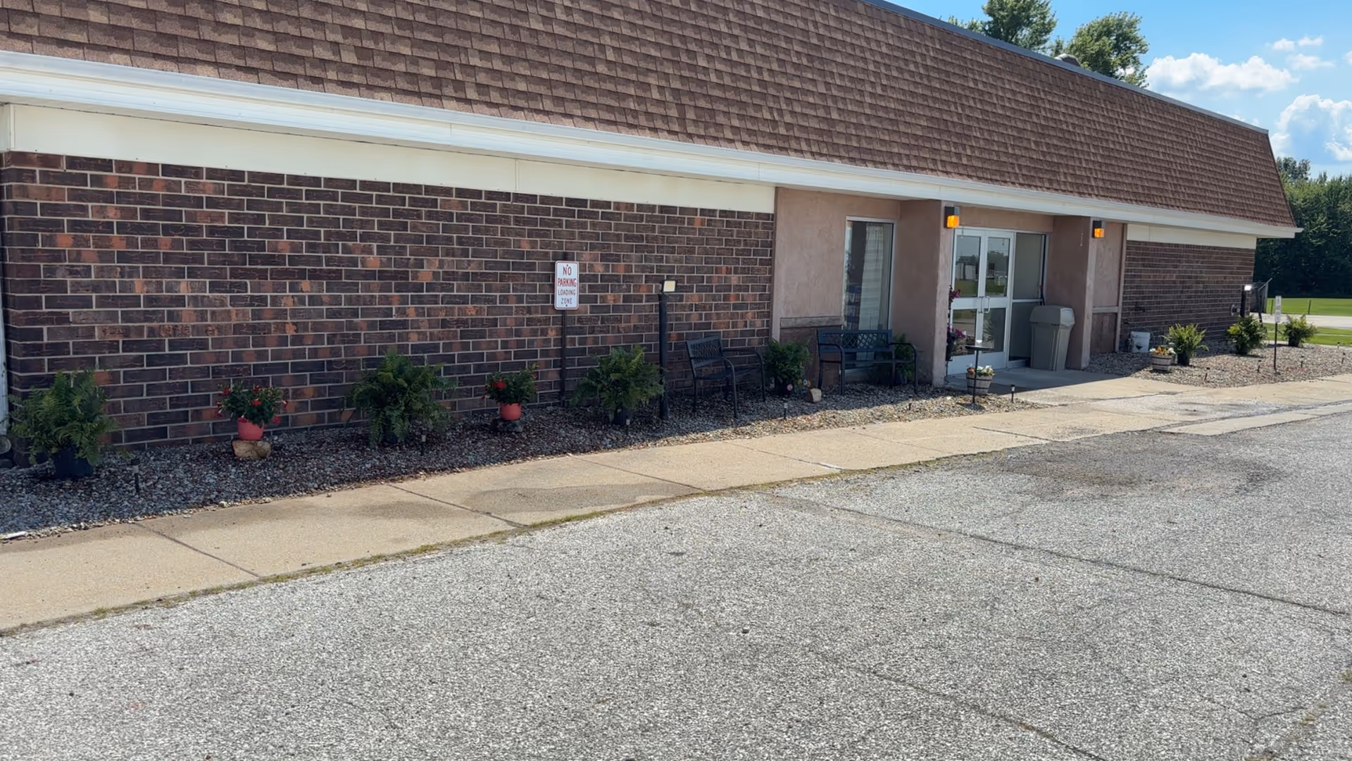 Exterior view of a single-story brick building with a brown shingled roof. There are several potted plants and small bushes along the front wall, two black benches near the entrance, and a sidewalk leading to a glass door entrance. The sky is clear and blue with some trees visible in the background.
