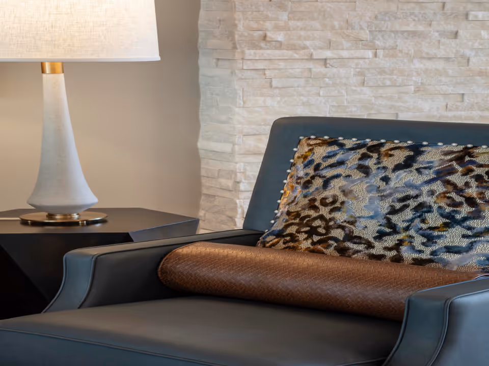 Close-up of a modern dark gray armchair with a textured brown bolster pillow and a leopard print decorative pillow, next to a black side table with a white lamp, set against a light stone textured wall.