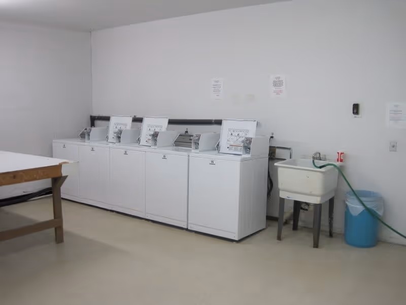 A laundry room with five white top-loading washing machines lined up against a white wall. To the right of the machines is a white utility sink with a green hose attached, and a blue trash bin next to it. On the left side, there is a wooden table with a white surface. The floor is light-colored and the walls are plain white.