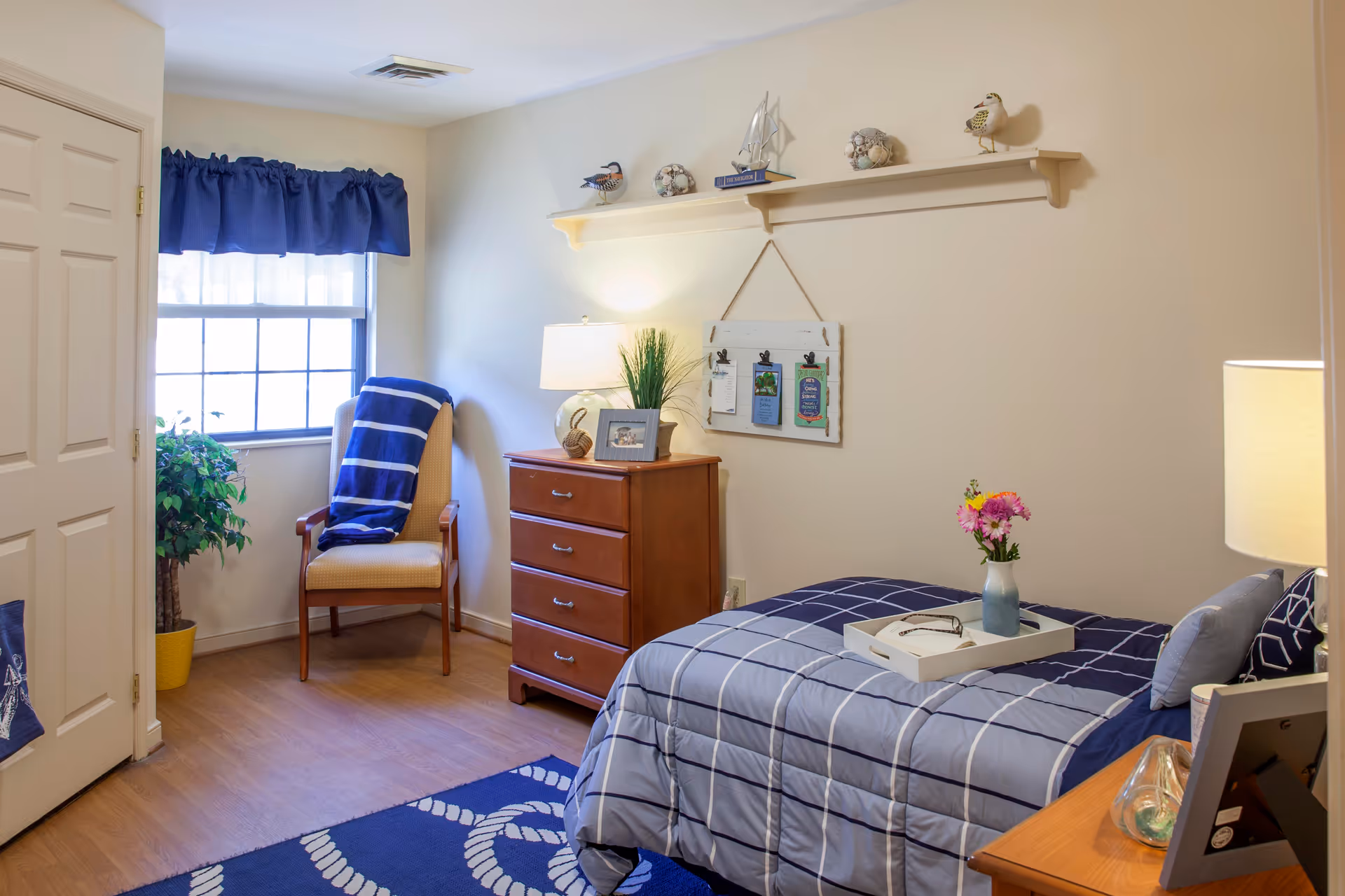 A cozy bedroom with a single bed covered in a blue and white checkered comforter. A white tray with a vase of flowers and a pair of glasses rests on the bed. Next to the bed is a wooden nightstand with a lamp and a decorative glass object. Across from the bed is a wooden dresser with a lamp, a framed photo, and a plant. A wooden chair with a blue and white striped blanket is placed near a window with blue curtains. The walls are decorated with a shelf holding small decorative items and a hanging board with clips. The floor has a blue rug with a white rope pattern.