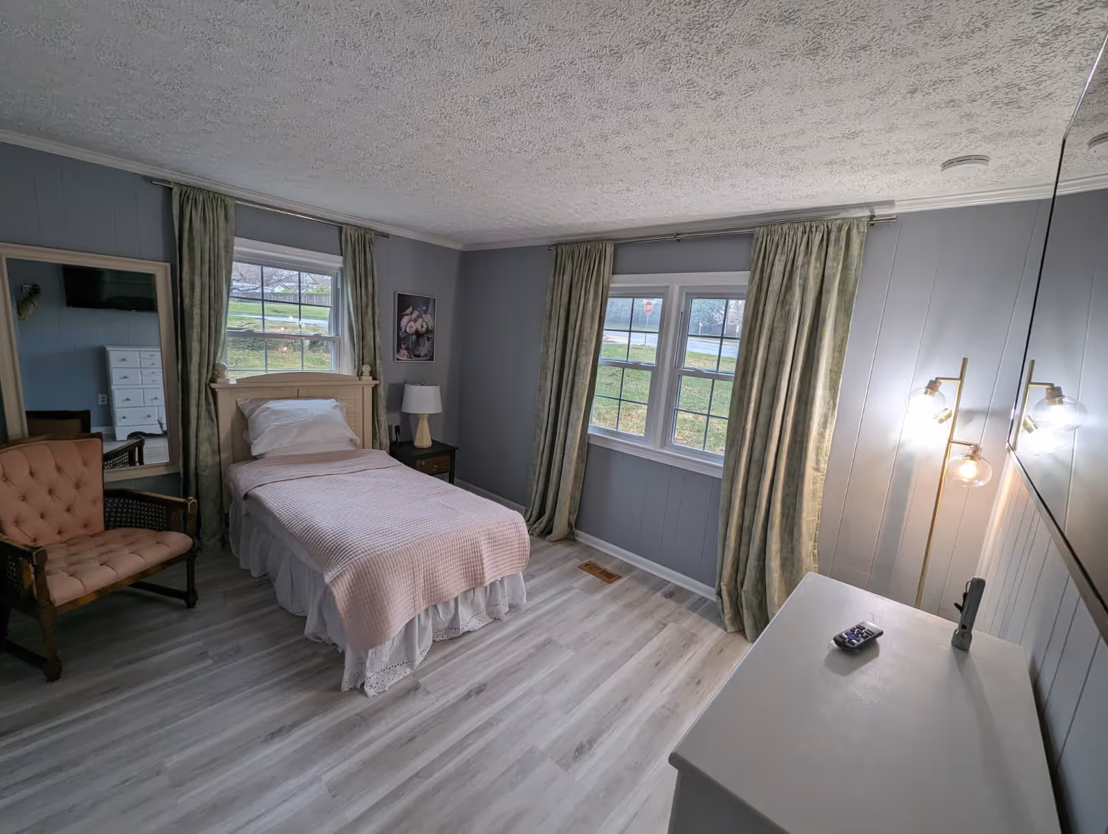 A cozy bedroom with a single bed, dresser, chair, and large windows with curtains.