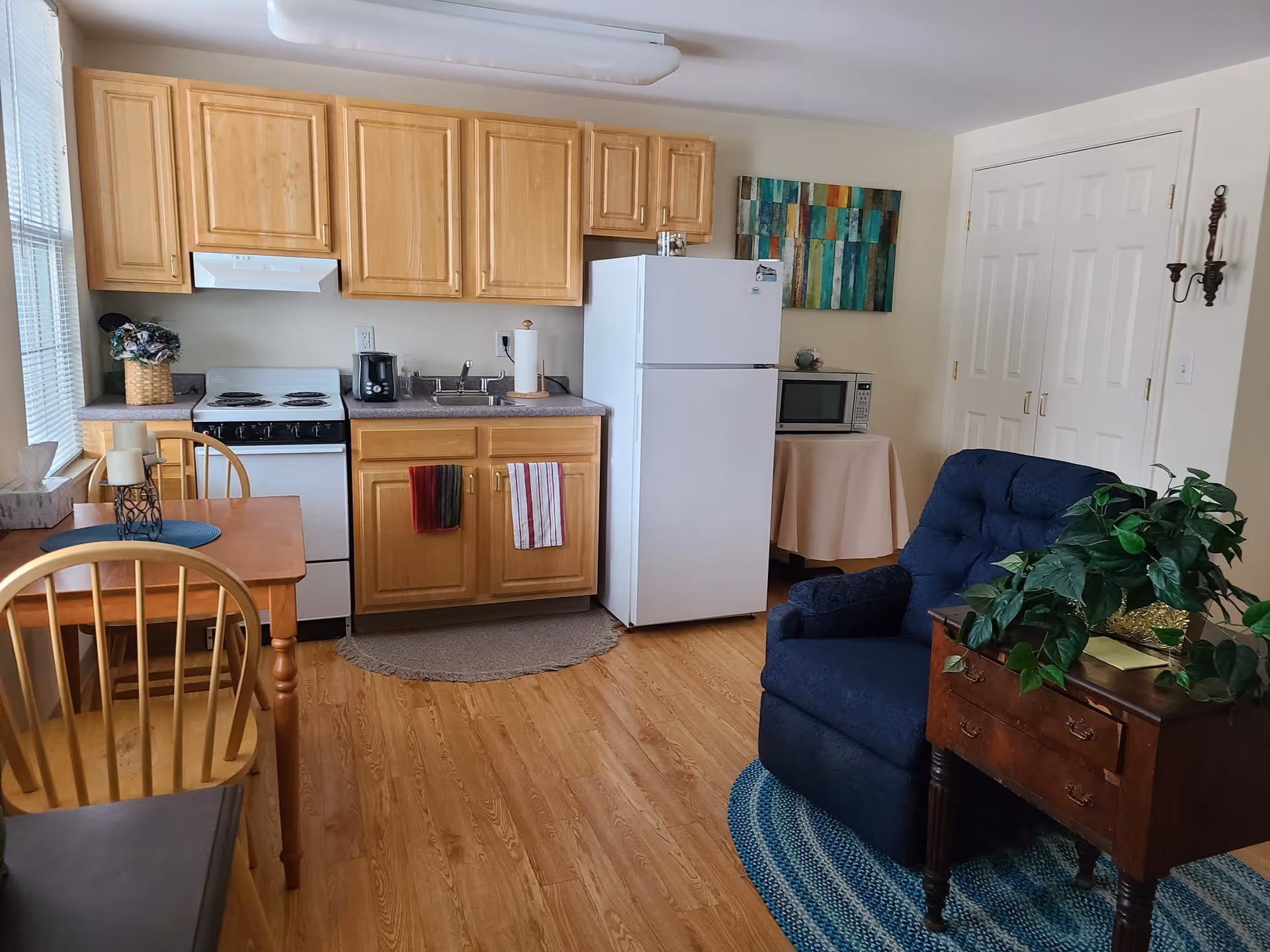 A small kitchen area with light wood cabinets, a white refrigerator, stove, and microwave on a table covered with a beige cloth. There is a wooden dining table with two chairs, a blue cushioned armchair on a blue braided rug, and a wooden side table with a green leafy plant. A colorful abstract painting hangs on the wall above the microwave.