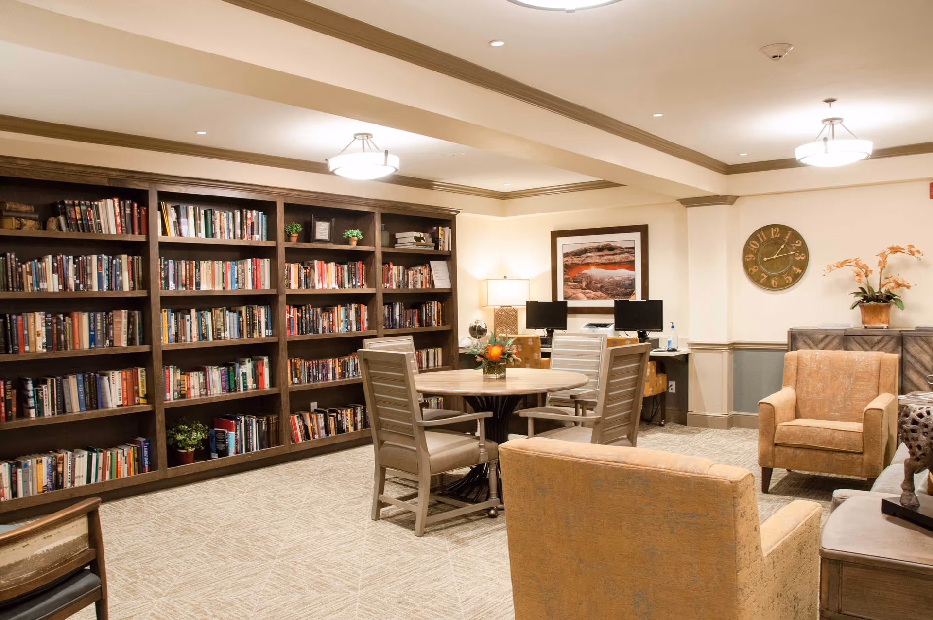 A cozy library and sitting area in a senior living facility with a large wooden bookshelf filled with books along one wall, a round table with four chairs in the center, two comfortable armchairs, a wall clock, a framed landscape picture, and two computer monitors on a desk in the background.