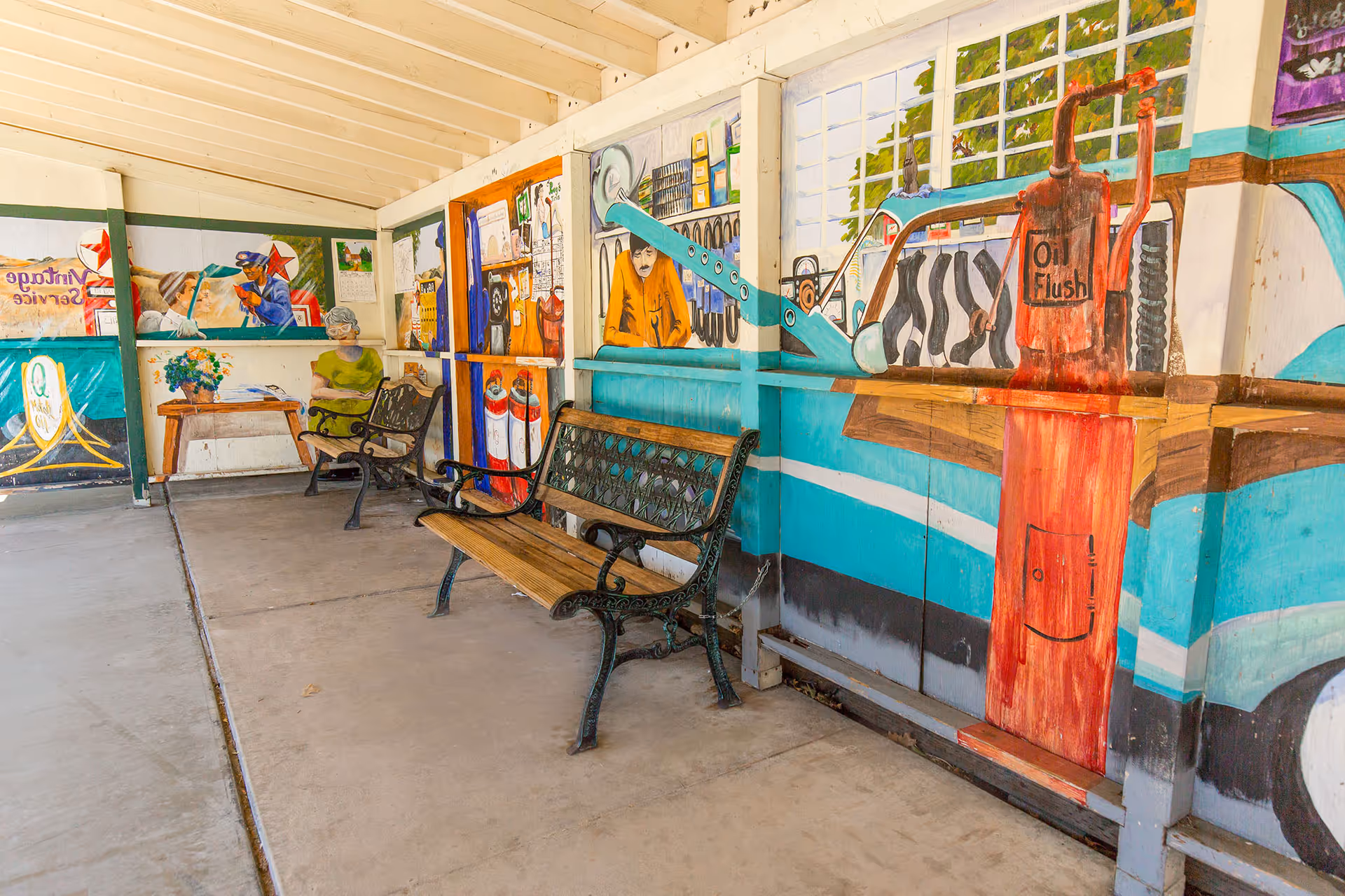 Covered outdoor seating area with metal and wooden benches. The walls are decorated with colorful murals depicting vintage service station scenes, including a gas pump labeled 'Oil Flush' and a man working on a car engine.
