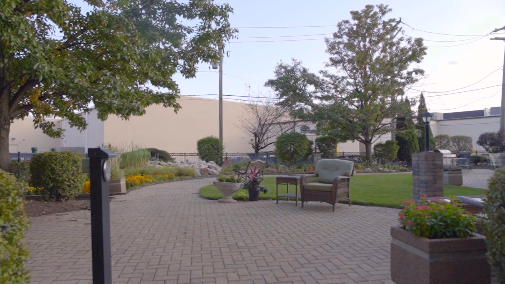 A paved outdoor garden area with green grass, trees, shrubs, and flower beds. There is a cushioned chair and a small table placed on the paved pathway. The background shows a beige building wall and some utility poles with wires.