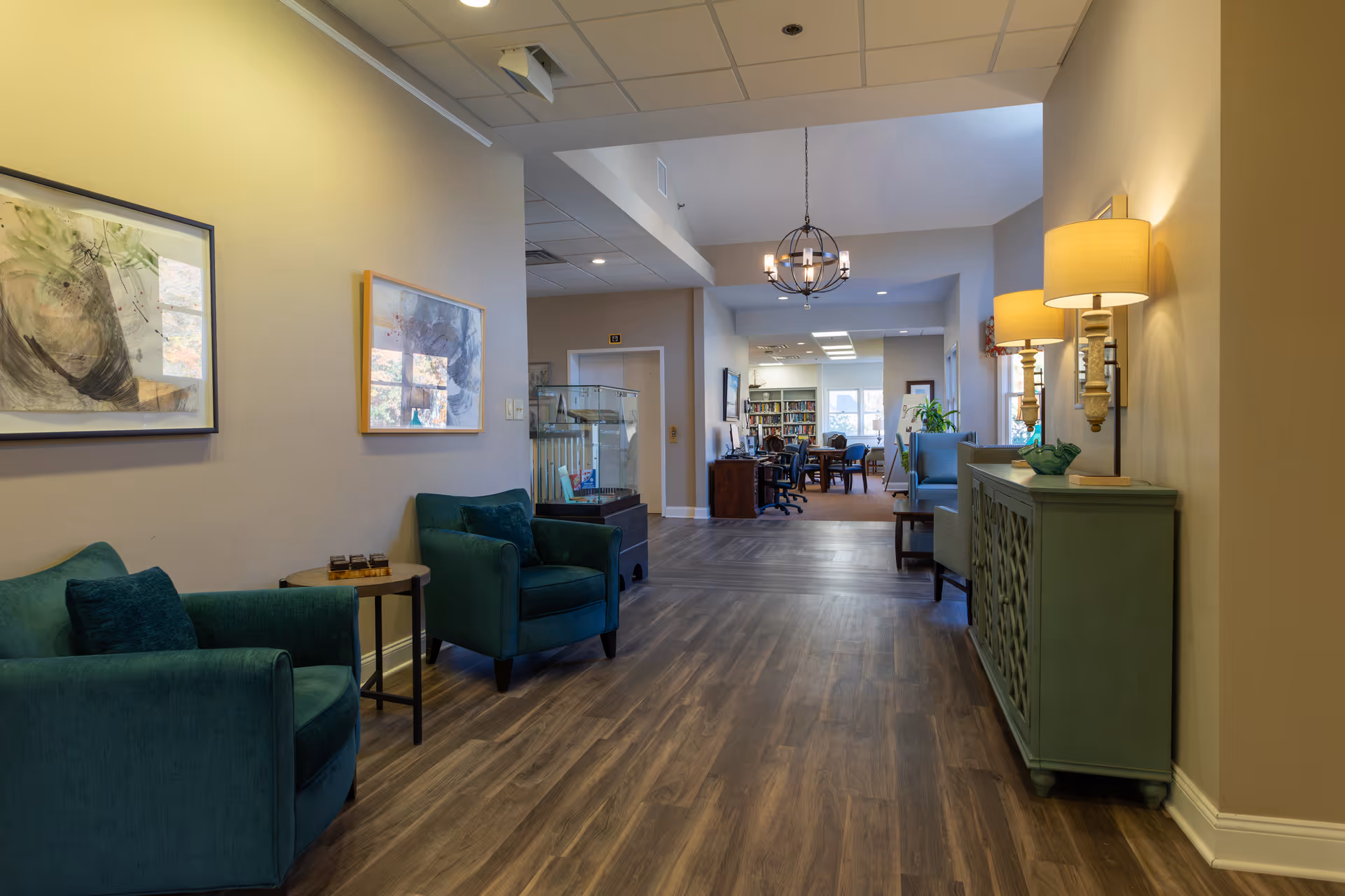 A well-lit hallway in a senior living facility with wooden flooring, two teal armchairs and a small round table on the left, and a green sideboard with two lamps on the right. The hallway leads to a room with bookshelves, chairs, and tables, suggesting a common area or library space. Abstract artwork is hung on the walls.