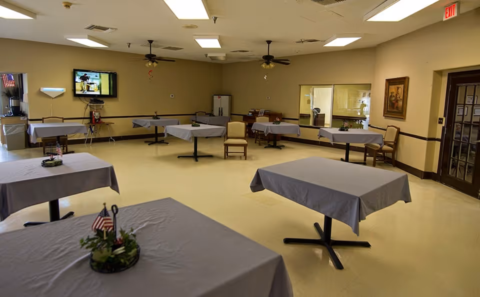 A dining room with several tables covered in gray tablecloths, each decorated with small centerpieces featuring miniature American flags. The room has beige walls, ceiling fans, fluorescent lighting, a wall-mounted TV, and a few chairs placed around the perimeter. There is an exit sign above a door on the right side of the room.