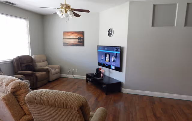 A senior living facility common area with three recliner chairs arranged around a wall-mounted flat screen TV. The room has wood flooring, a ceiling fan with lights, a window with blinds, a wall clock, and a landscape painting on the wall.