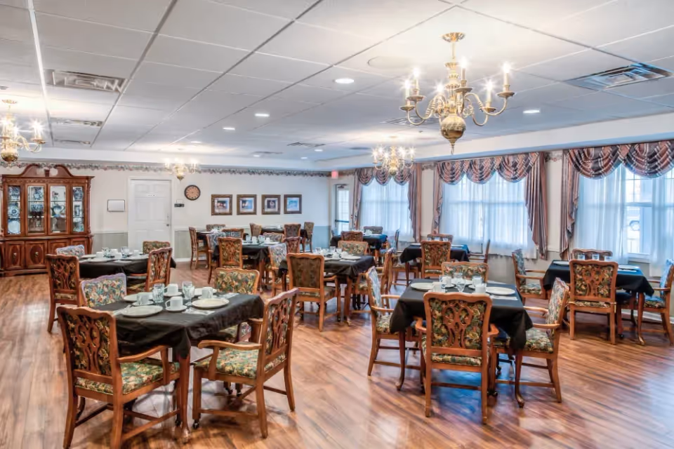 A spacious dining room with multiple wooden tables covered with black tablecloths, each set with white plates, cups, and silverware. The room features wooden chairs with floral upholstery, large windows with decorative curtains, chandeliers hanging from the ceiling, and a wooden china cabinet against one wall.