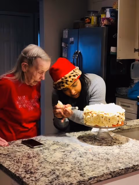 An elderly woman in a red sweater and a younger woman wearing a red and leopard print Santa hat are decorating a white frosted cake together on a kitchen counter. The kitchen has a granite countertop, a refrigerator, and cabinets in the background.