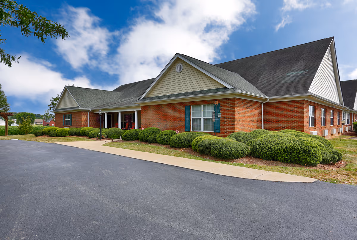 Brick ranch-style building with a covered entrance, trimmed shrubs, and a paved driveway under a partly cloudy sky.