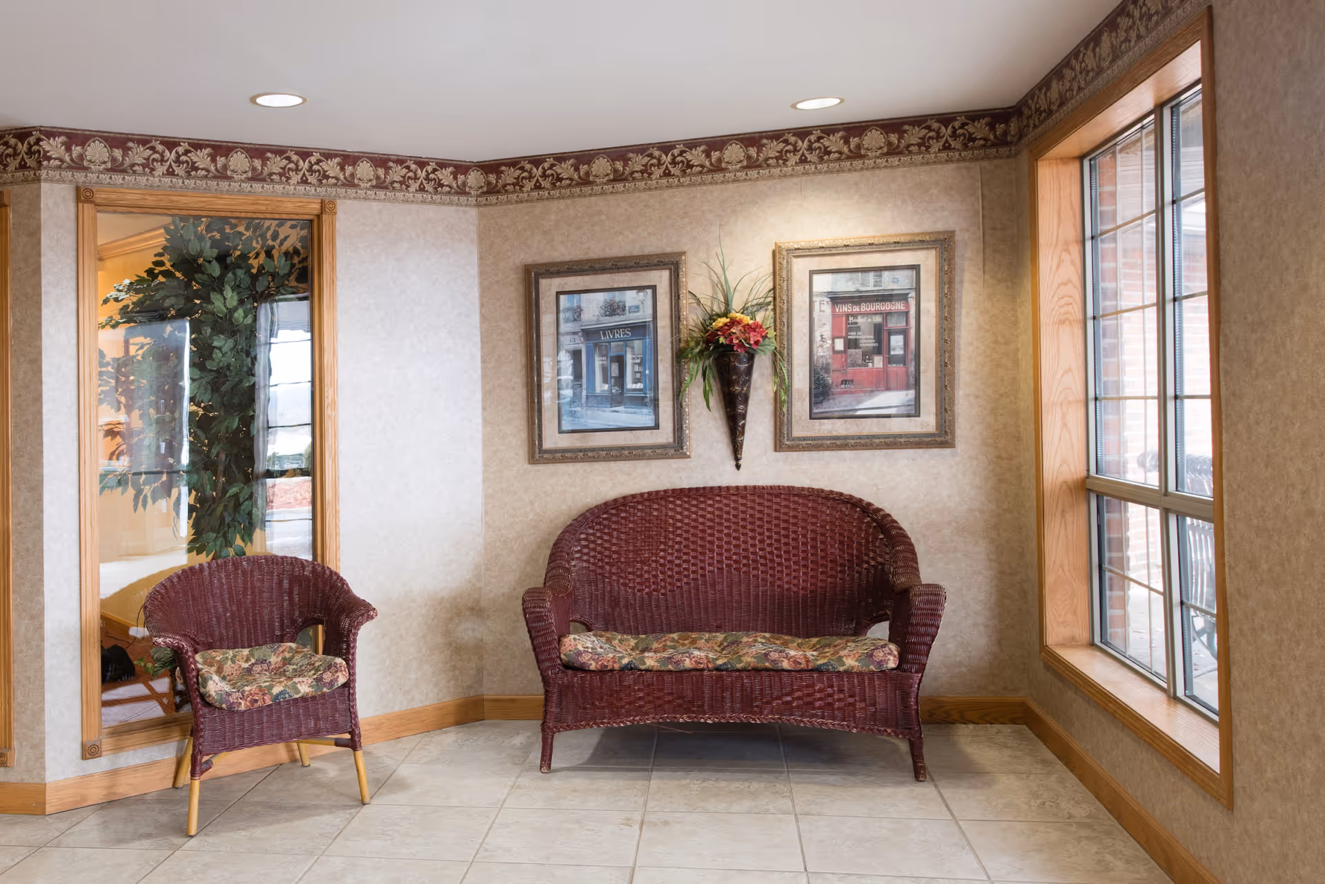 A cozy seating area with a maroon wicker loveseat and matching chair, both with floral cushions. The walls are decorated with two framed pictures and a wall-mounted floral arrangement. There is a large window with wooden trim letting in natural light, and a mirror reflecting a plant and part of another room.