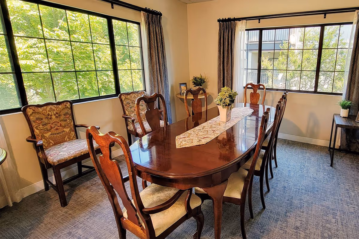 Sunlit dining room with a polished wooden table and upholstered chairs beside large windows overlooking trees.