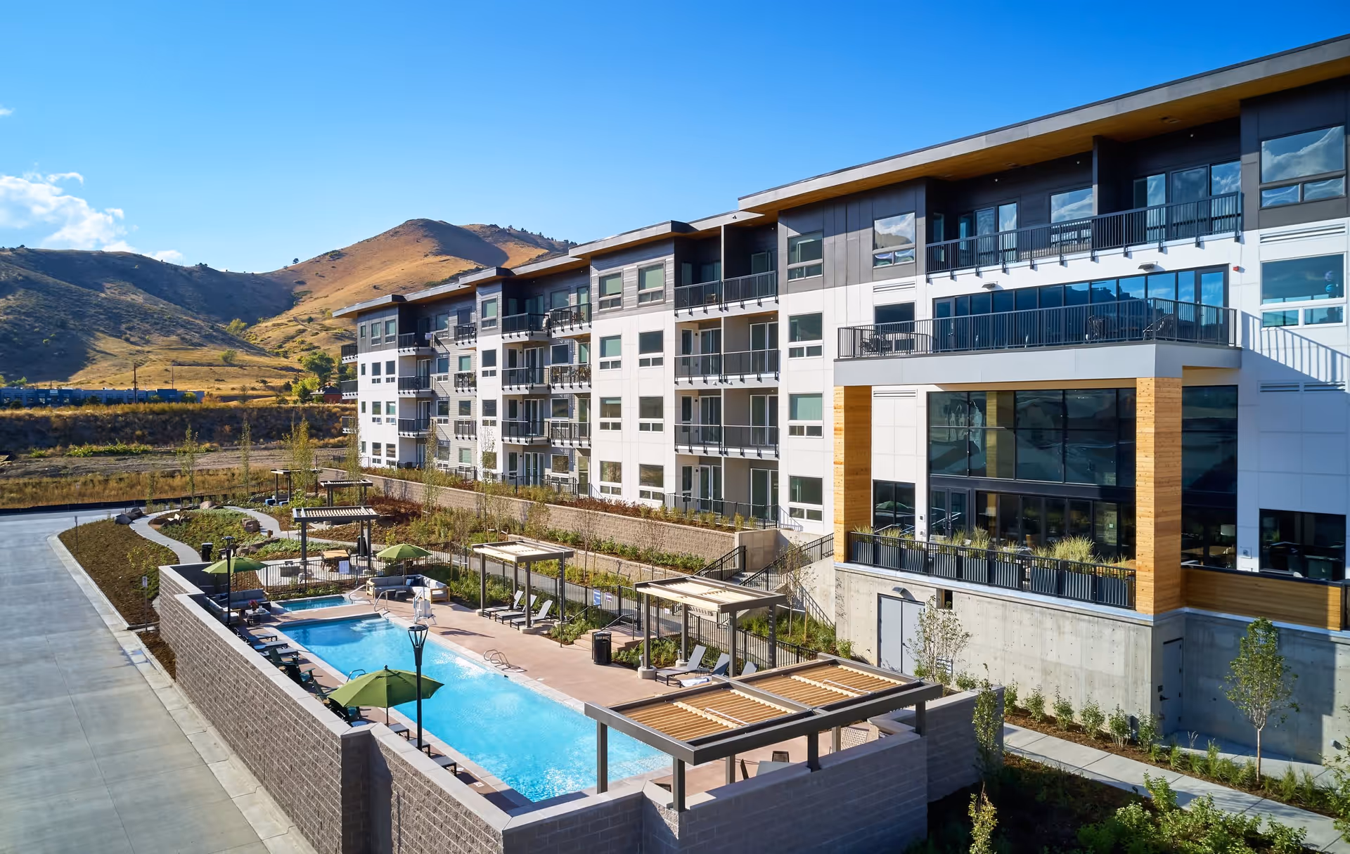 Exterior view of a modern multi-story senior living building with balconies, an outdoor pool and pergolas set against rolling hills.