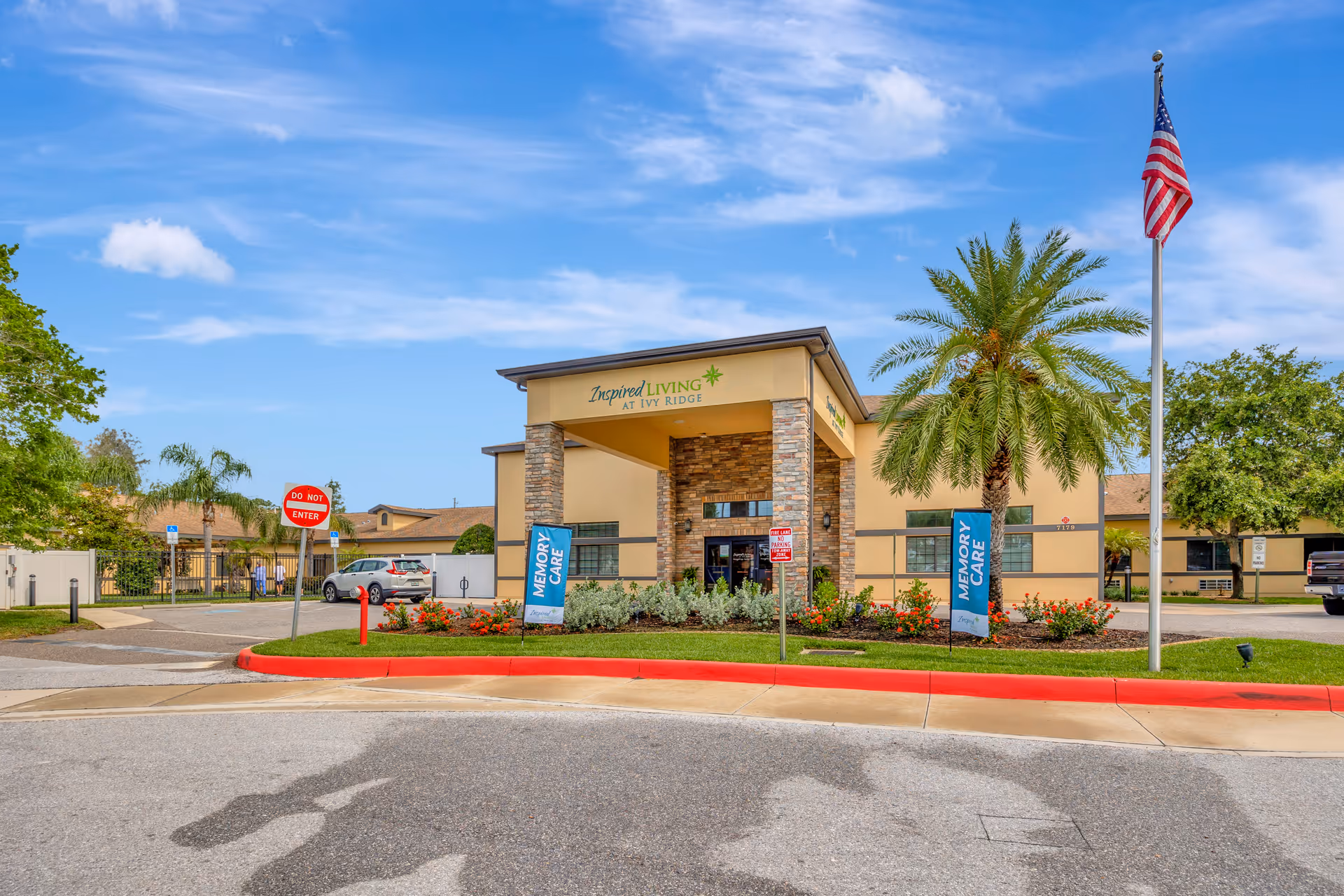 Front exterior view of Inspired Living at Ivy Ridge facility with a covered entrance, stone pillars, landscaped flower beds, palm trees, an American flag on a flagpole, and a clear blue sky.